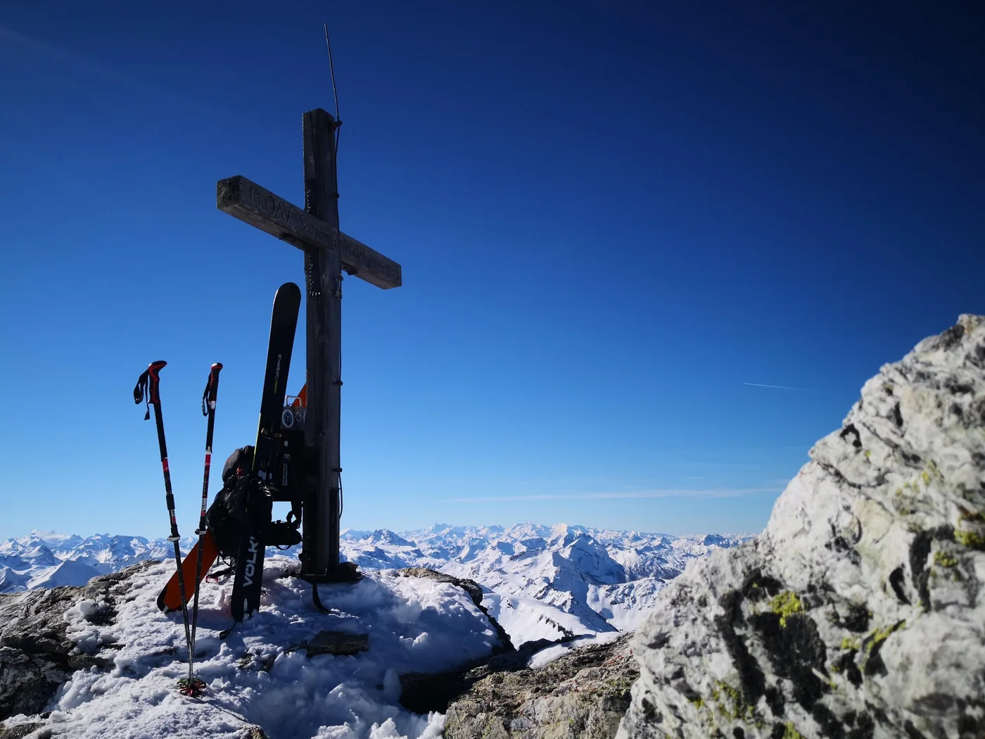 Ski und Stöcke am Gipfelkreuz vor einem weiten Panorama verschneiter Berge