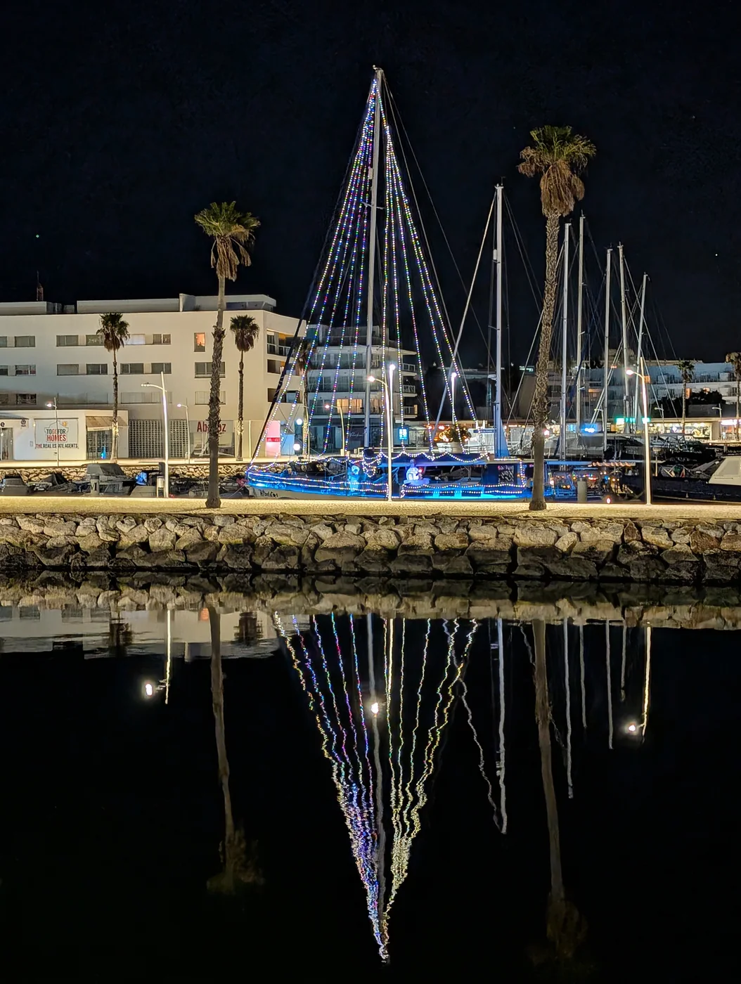 Lichter an einem Segelmast spiegeln sich nachts im Hafenwasser